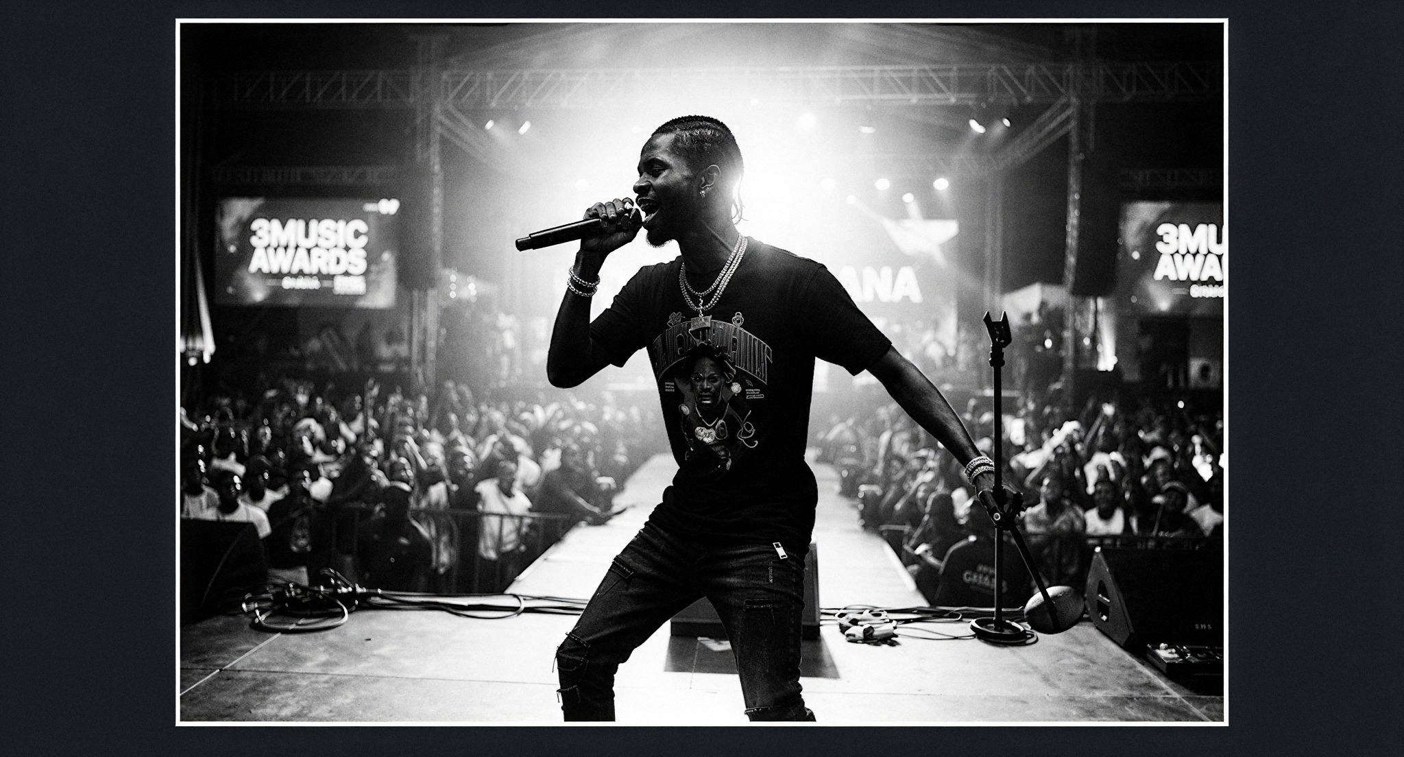 A high-contrast black and white portrait of the Ghanaian artist Black Sherif performing on a brightly lit stage during the 3Music Awards. The lighting creates a dramatic halo effect around his silhouette. The style is raw and impactful, capturing a historic cultural moment of discovery and artistic power. The image is framed with a 1px electric white border.