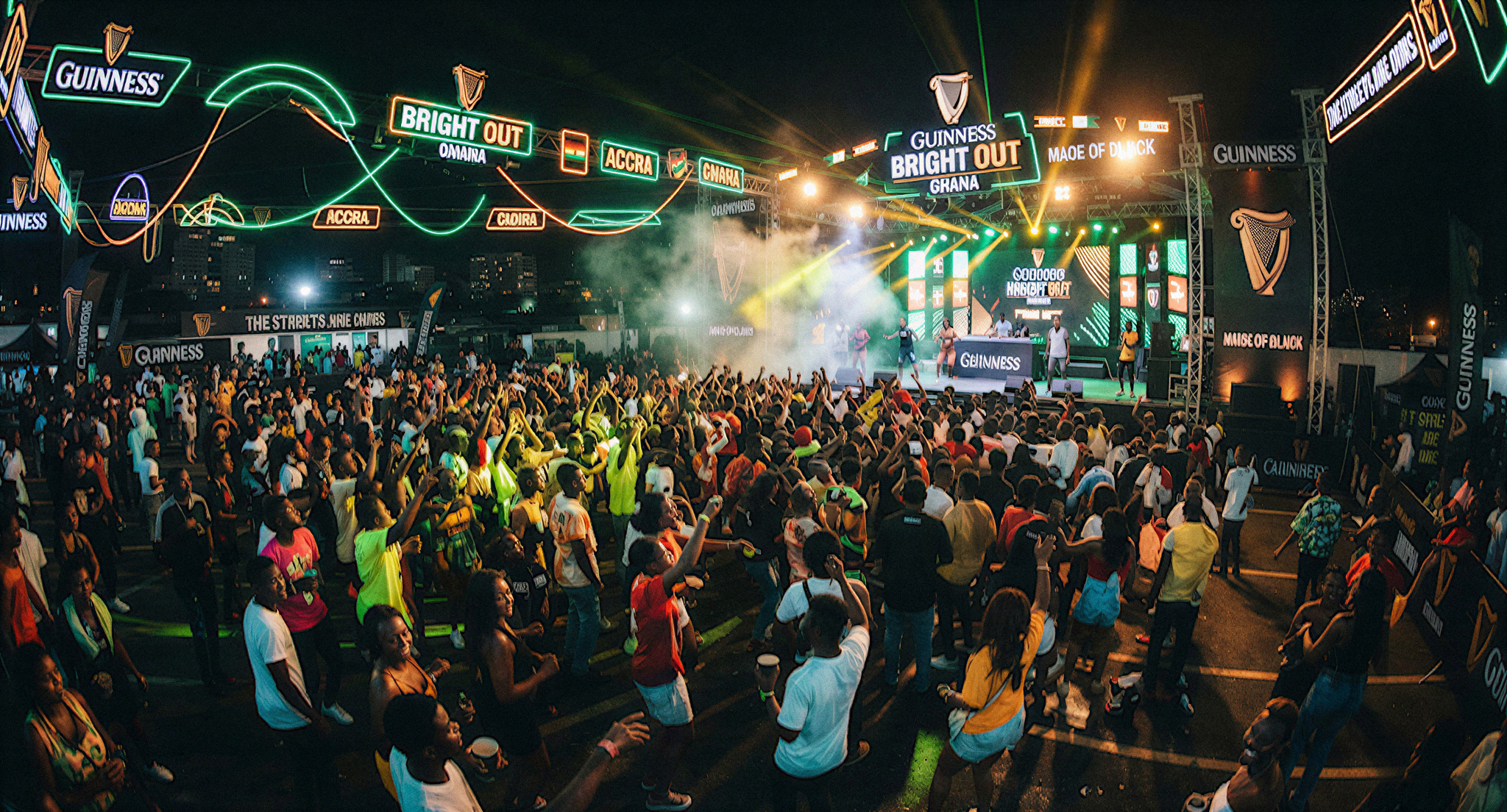 A vibrant, wide-angle shot of a Guinness Bright Out festival activation in Ghana. The scene is packed with energetic youth dancing under glowing neon green and amber lights. Large Guinness branding and custom stage designs dominate the space. The lighting is electric and festive, capturing a high-energy cultural moment with rich, saturated colors and deep shadows.