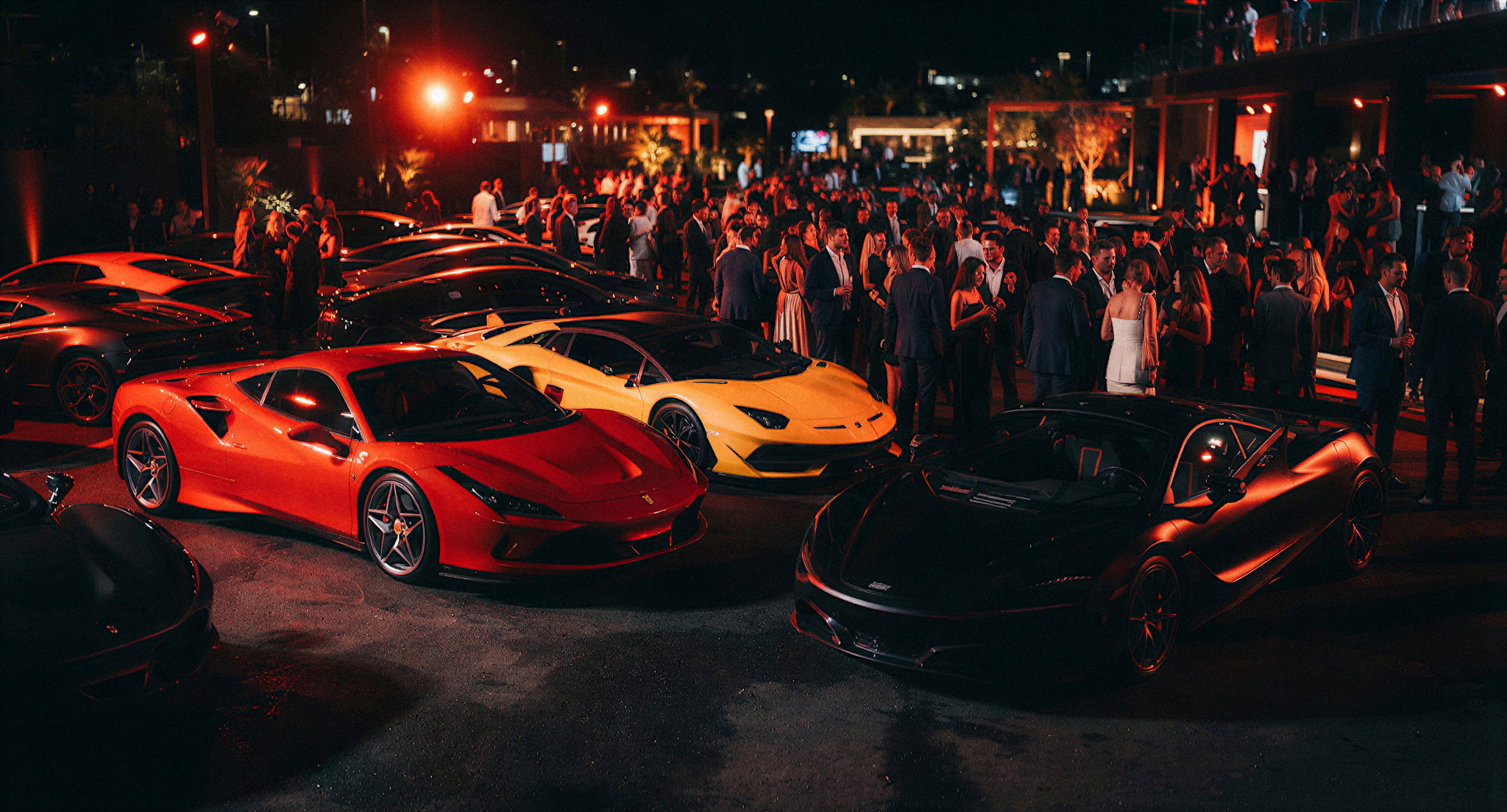 A high-energy, cinematic shot of a luxury supercar event at night. Sleek Ferraris and Lamborghinis are parked under dramatic red and orange spotlights. A large crowd of stylishly dressed attendees is visible in the background, creating a sense of exclusivity and motion. The aesthetic is high-contrast, moody, and premium, dominated by deep blacks and vibrant red lighting.