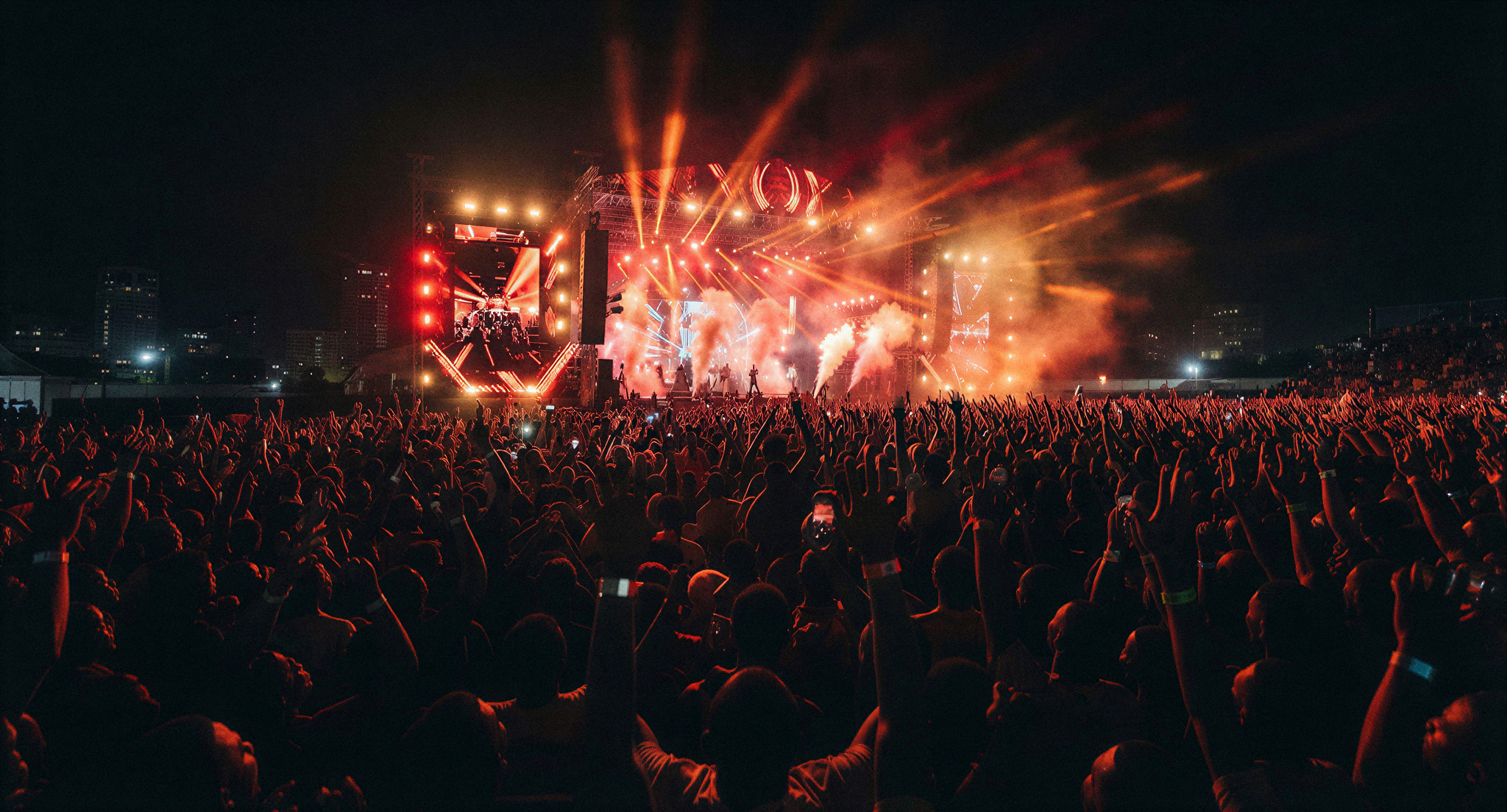 A cinematic, low-angle photograph of a massive outdoor music festival stage at night in Accra, Ghana. Thousands of silhouettes of hands are raised against a backdrop of vibrant red and orange laser lights. The atmosphere is electric and high-energy, with a thin veil of stage smoke catching the light. The photography is editorial and professional, emphasizing the sheer scale of the 3,200 person crowd.